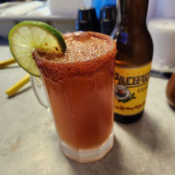 A michelada with a lime wedge and chili-salted rim, next to a bottle of Pacifico beer.