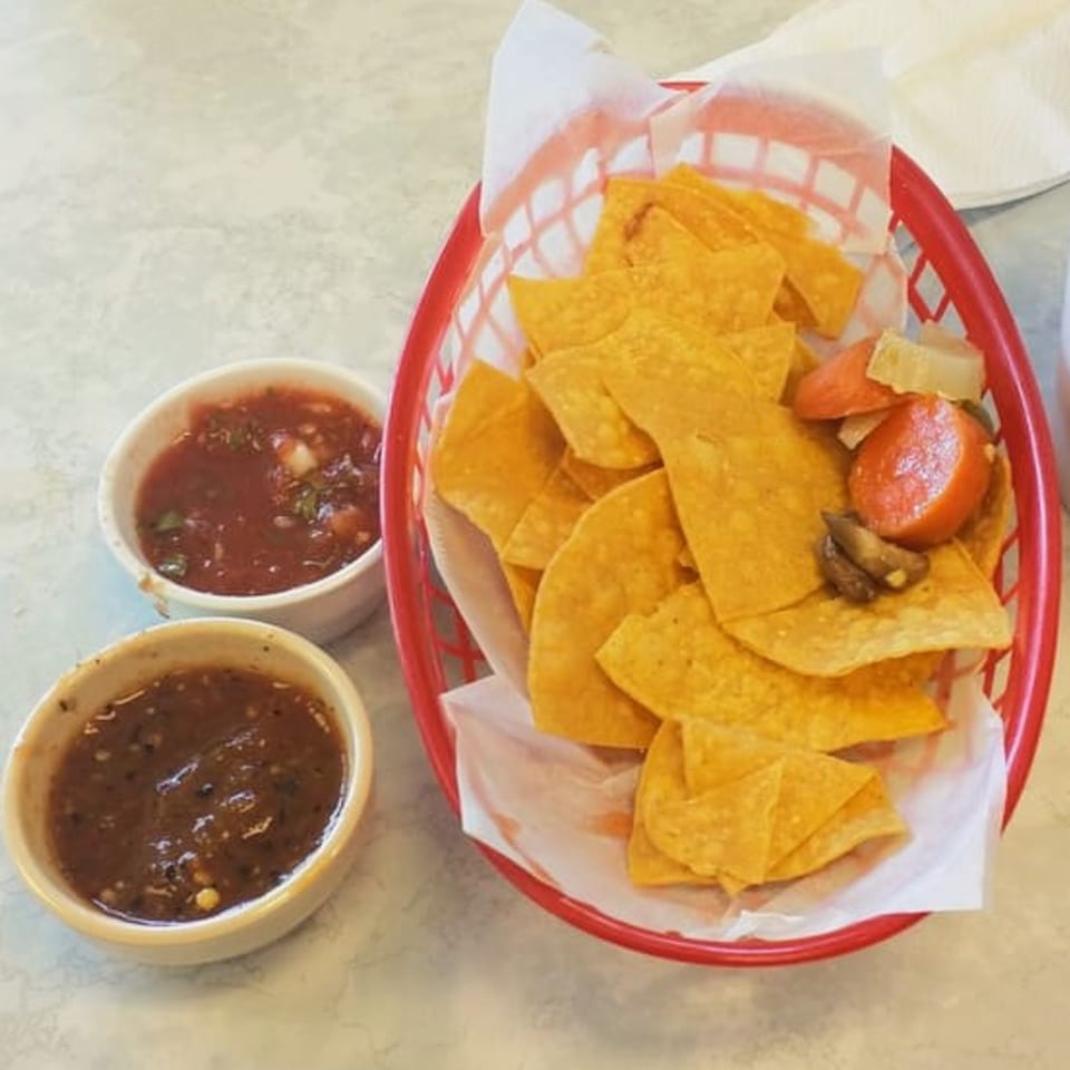 A red basket of tortilla chips with two small bowls of salsa on a light-colored table.