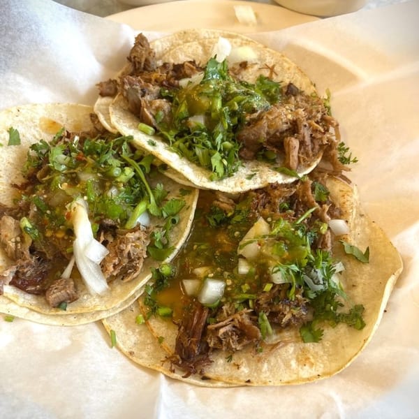 Three carne asada street tacos with green salsa, cilantro, and onions on white paper.